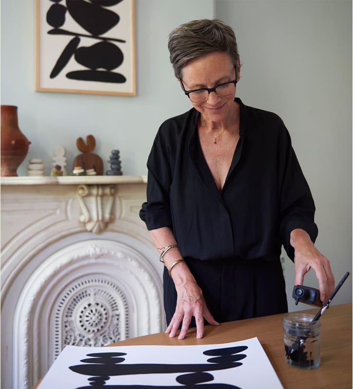 A woman paints black brushstrokes on white paper in a stylish room.